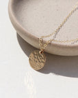 14k gold fill Moon drop necklace laid on a plate in the sunlight. This necklace features our simple chain met by a hammered coin, making it look like the moon.