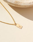 A gold rolo chain with a stamped mini tag with the letter "B". Necklace is displayed next to a stone dish.