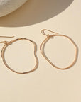 A gold large hoop earrings, each uniquely shaped, hang elegantly from simple hooks. Earrings are displayed on a cream backdrop.