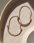 14k gold fill hammered Paloma hoops, earrings are placed on a peach colored plate in the sunlight. These earrings feature a wire around 1/2 inch shaped like an oval, then followed by the other half of the oval that is hammered.