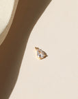 Gold charm featuring a clear teardrop-cut crystal, photographed on a warm neutral surface with soft natural light and shadows.