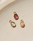 Three oval gemstone charms in pink, green, and peach tones set in gold bezels, displayed on a neutral textured surface in warm natural light.
