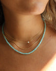 Model wearing a styled necklace stack. Featuring a simple 14k gold fill chain with a mini pearl anchored in the center. It is paired with a rolo 14k gold fill chain, and a fully beaded gemstone necklace to create a three necklace stack.