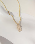 A gold rolo chain with a stamped mini tag with the letter "B". Necklace is displayed over a white dish.