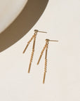 Two 14k gold fill chain stud earrings hanging off a small ball stud, the chain features a bar + link style. The earrings are displayed on a cream back drop in the sunlight.
