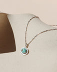 Silver paperclip-style chain necklace featuring a round turquoise stone with natural brown matrix, framed by a sunburst-textured bezel, resting on a speckled beige ceramic display under warm sunlight.