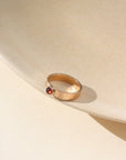 A ridged textured 14k gold fill band with a 4mm red garnet gemstone set in the middle. Ring is displayed on a tan dish.