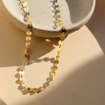 14k gold fill Starlight Choker laid in the sunlight on a gray plate. This choker is perfect for layering. This Necklace features circle disc that are linked together.