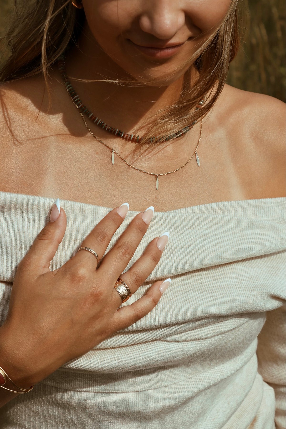 Close-up of layered gold necklaces, gold rings, and bracelets styled with a cream off-the-shoulder top in warm sunlight.
