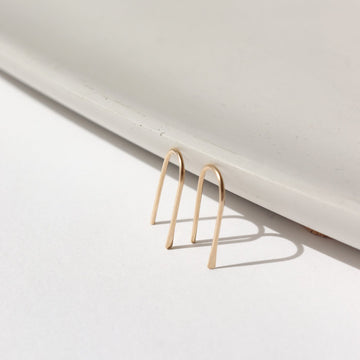 14k solid gold wire formed into arch shaped earrings with one hammered end, photographed on a white table leaning on a white dish