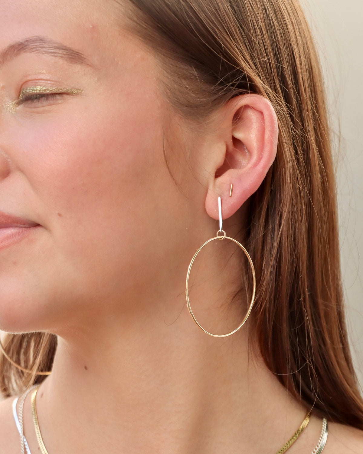 Close-up of a model wearing mixed-metal hoop earrings featuring silver bar studs and large gold circular drops, paired with layered necklaces.