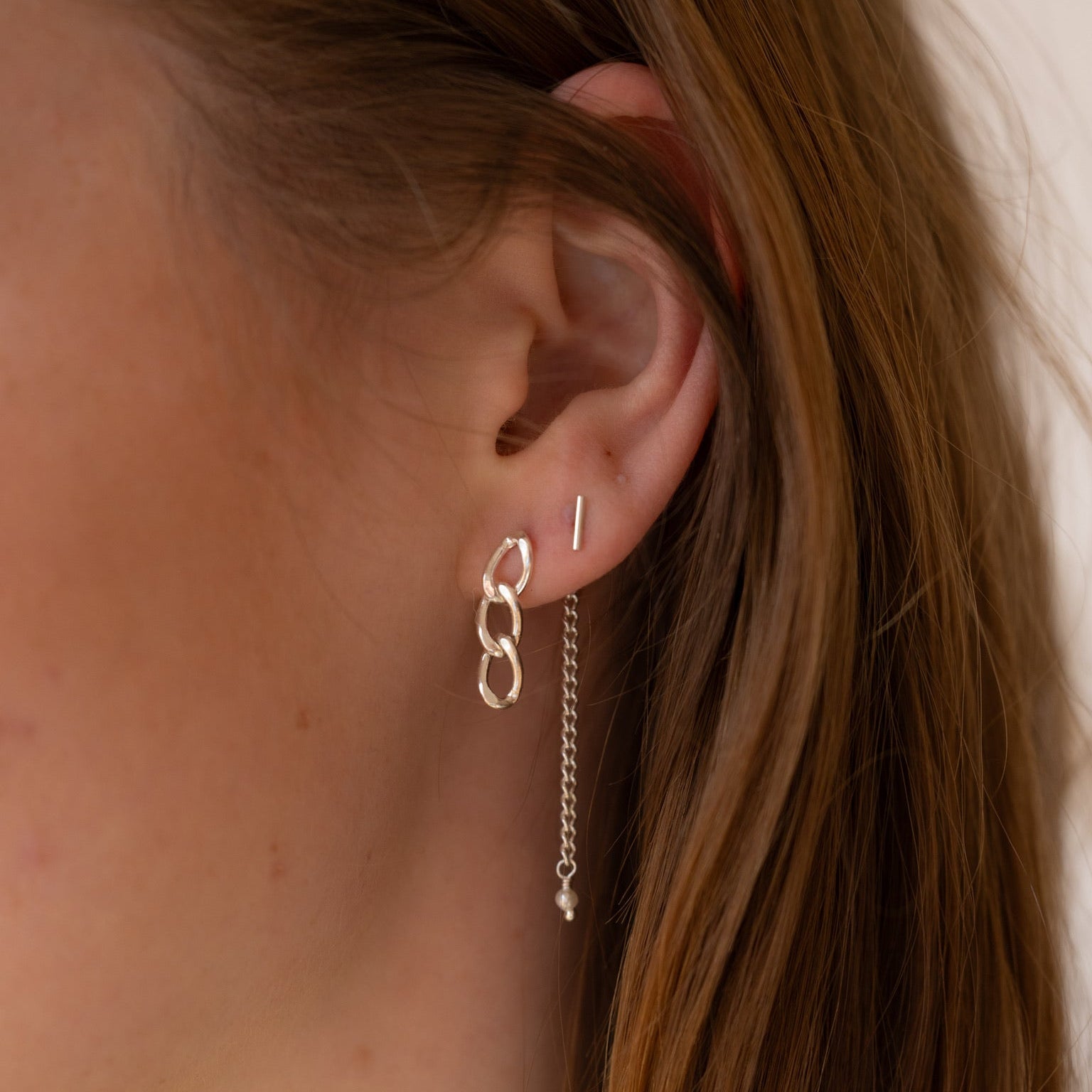 A close-up of a person's ear wearing a silver dangle earring with interlocking oval links and a short chain drop, alongside a simple vertical silver stud earring.