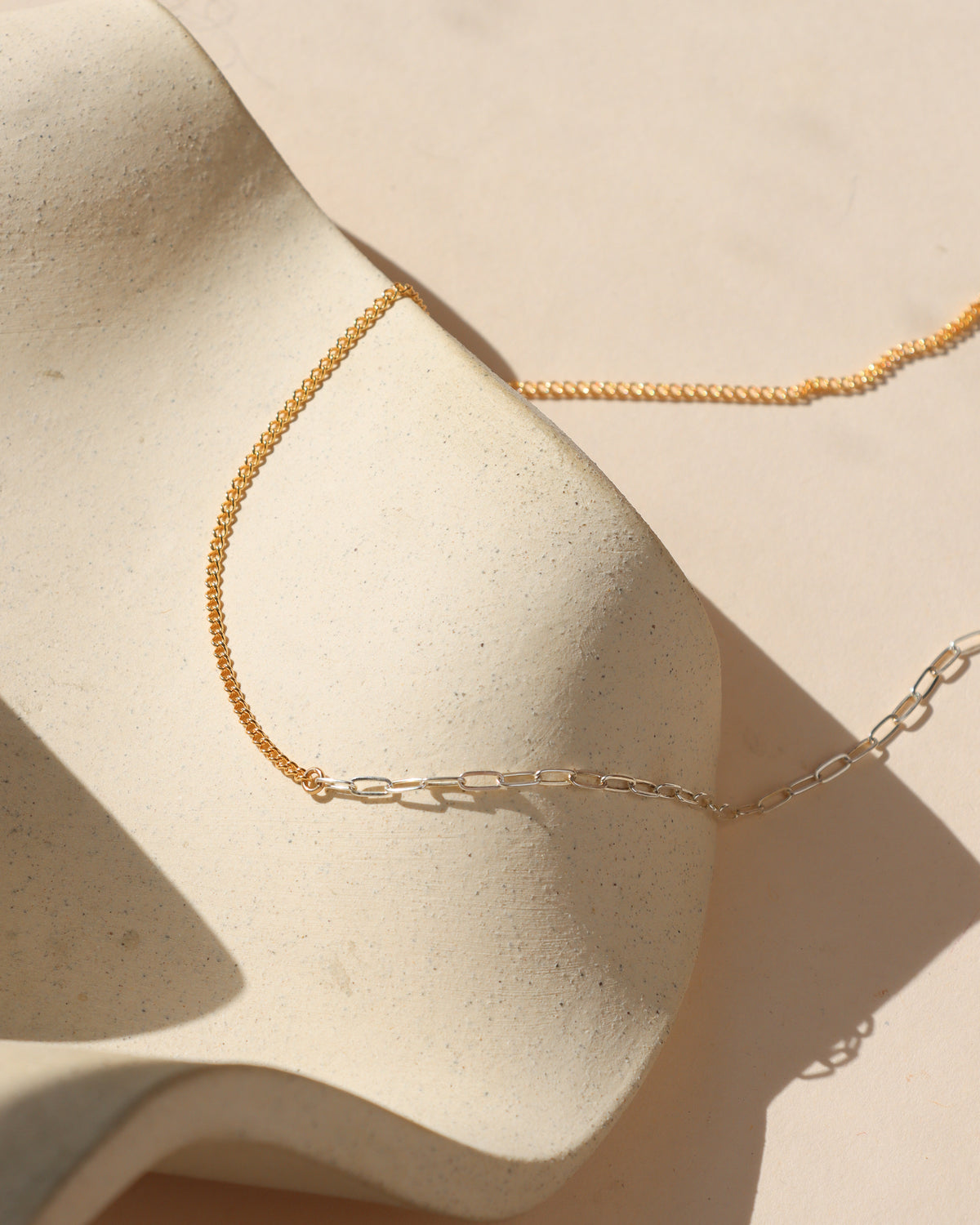 Balance Necklace featuring a delicate 14k gold fill chain joined with elongated sterling silver paperclip links, draped over a cream ceramic surface in sunlight.