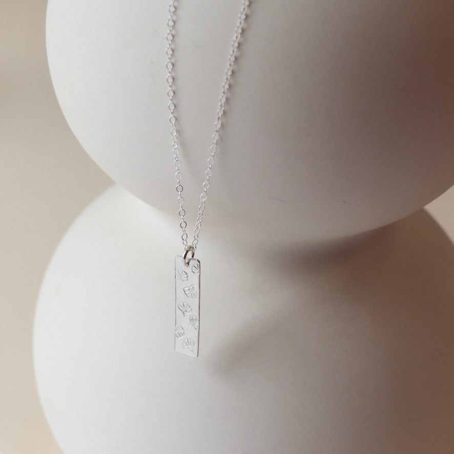 sterling silver delicate chain necklace with handmade sterling silver rectangle pendant with flowers stamped on it on a light dish