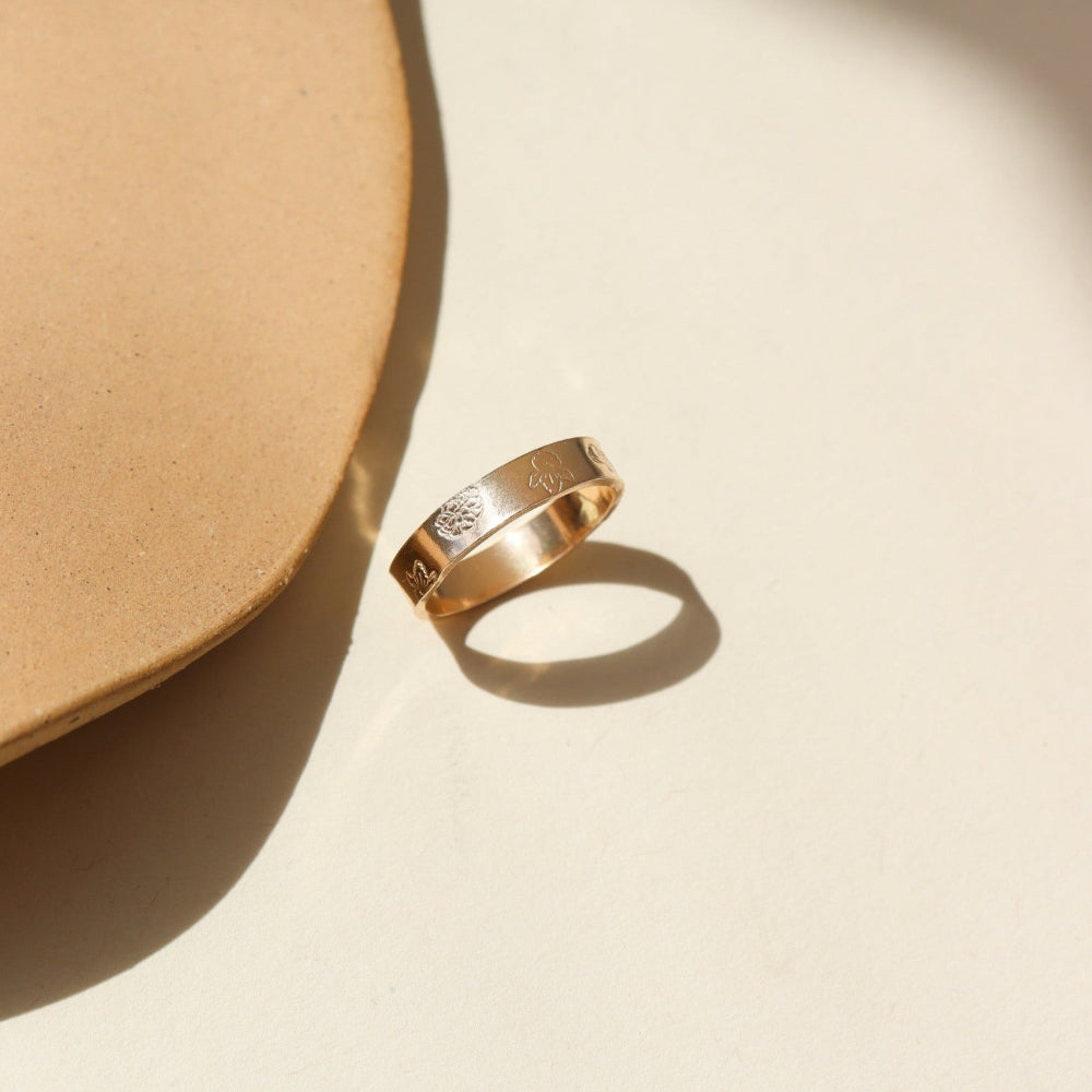 A thick 14k gold band filled with stamped birth flowers spread on the band. The ring is displayed on a cream back drop.