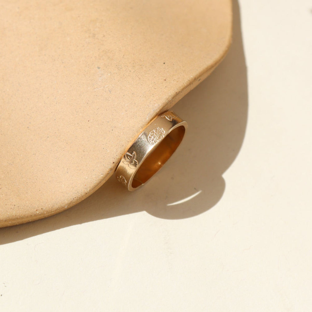 A thick 14k gold band filled with stamped birth flowers spread on the band. The ring is displayed on a cream back drop.