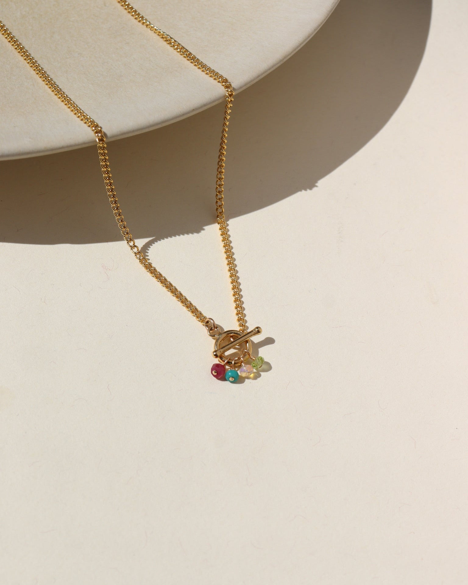A 14k gold fill toggle necklace featuring a curb chain with four gold wire wrapped birth stones dangling from the toggle. Necklace is displayed on a cream back drop. 