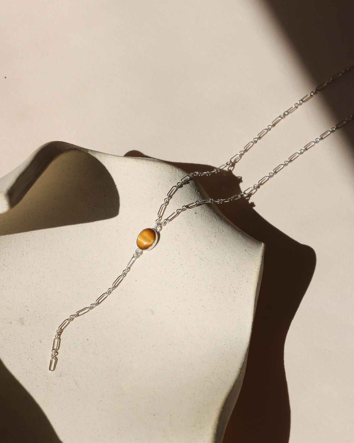 Silver Y-necklace featuring an oval tiger’s eye gemstone and paperclip chain drop, displayed on a neutral surface in sunlight.