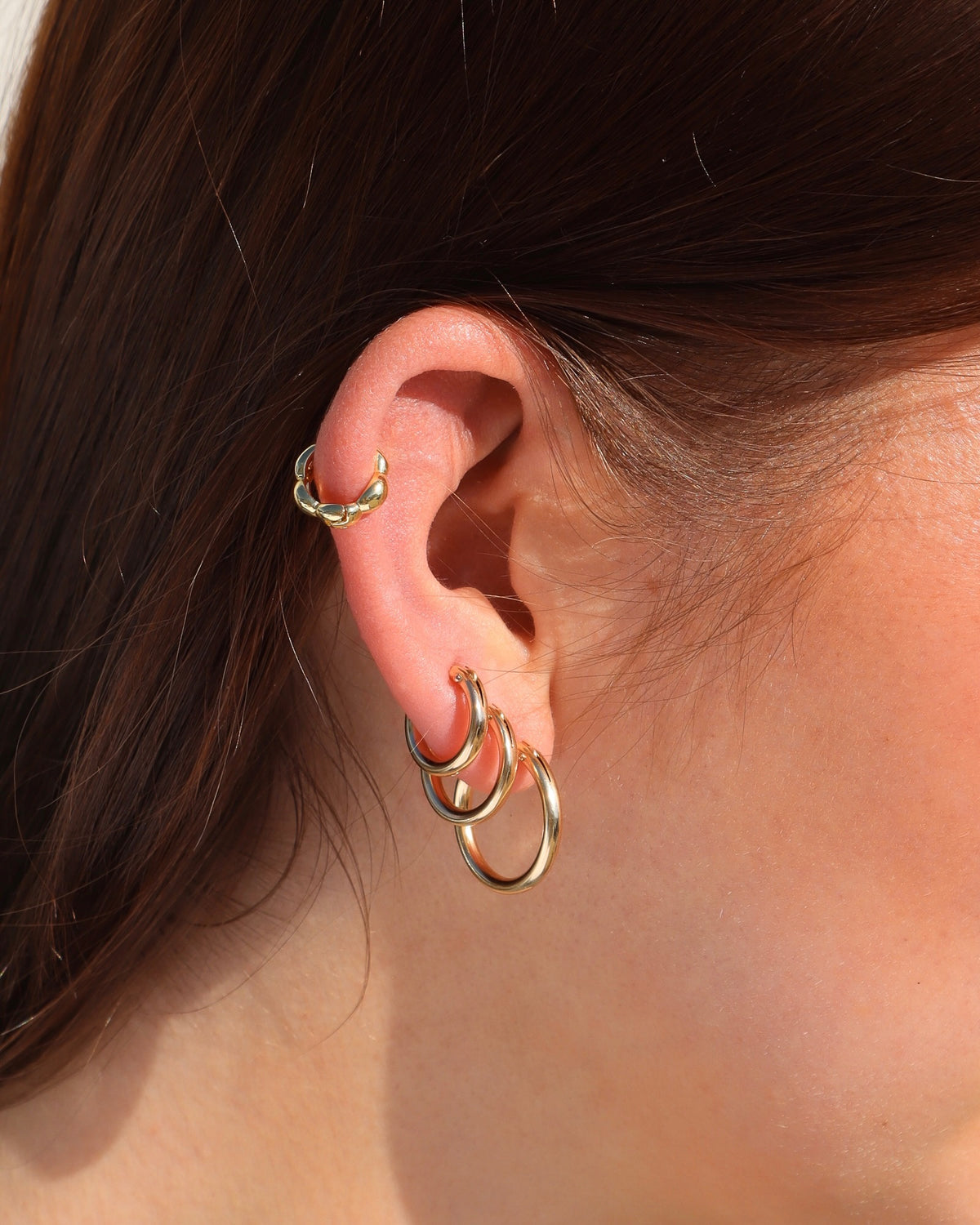 Close-up of an ear styled with three polished gold hoop earrings in graduating sizes and a sculptural gold hoop on the upper cartilage, creating a layered gold ear stack.