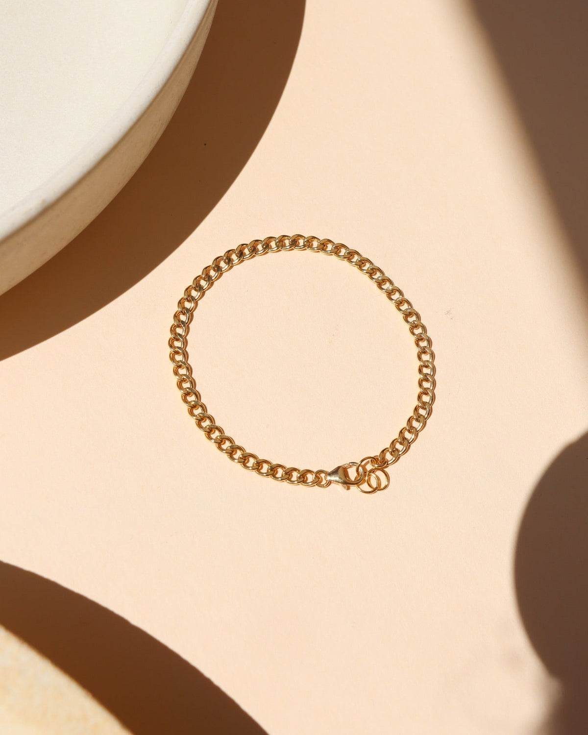Classic gold curb link chain bracelet featuring interlocking flattened links and a spring ring clasp, laid flat on a peach-beige surface near a ceramic dish with warm natural light shadows.