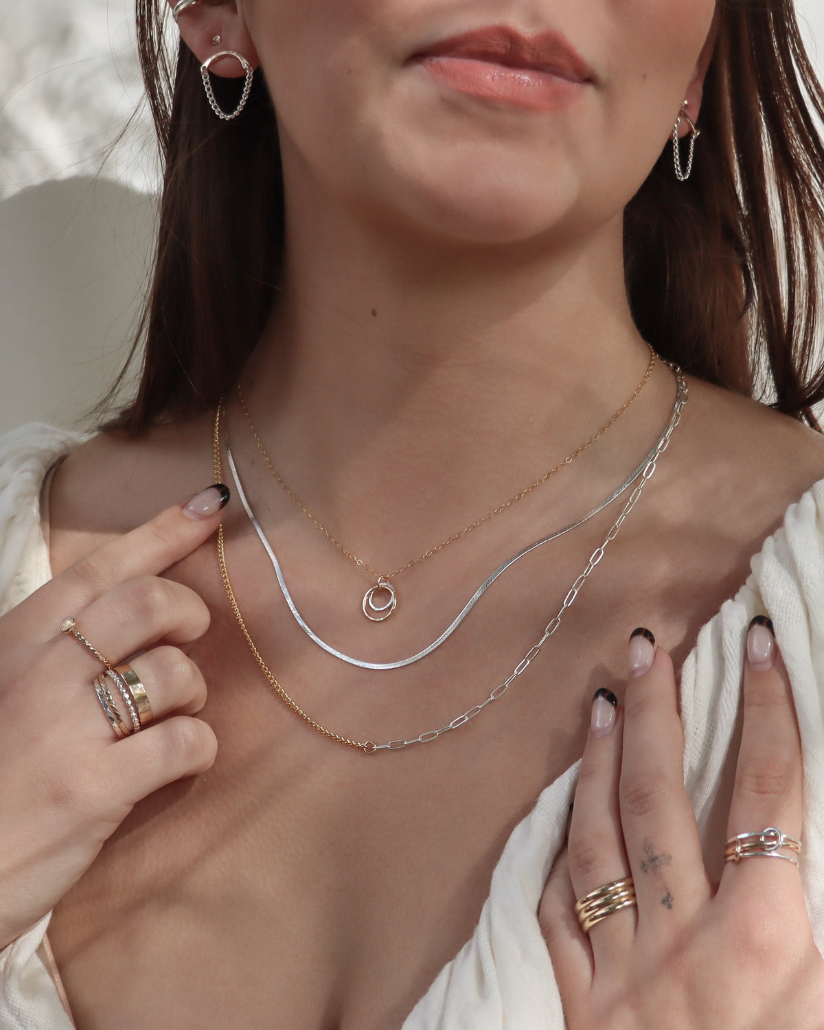 Model wearing layered gold and silver necklaces, including a mixed metal Balance Necklace, silver herringbone chain, and delicate circle pendant, paired with Chandelier Studs and mixed metal rings.