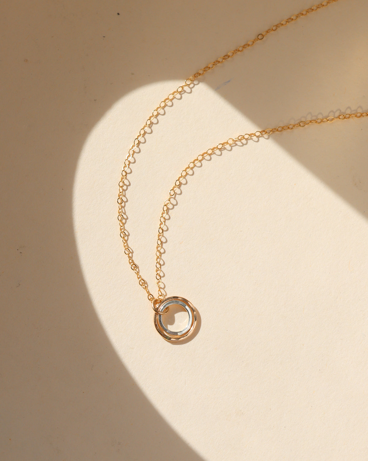 Close-up of a Token Jewelry mixed metal pendant necklace featuring interlocking gold and silver circular rings on a delicate gold chain, displayed on a cream background in natural sunlight.