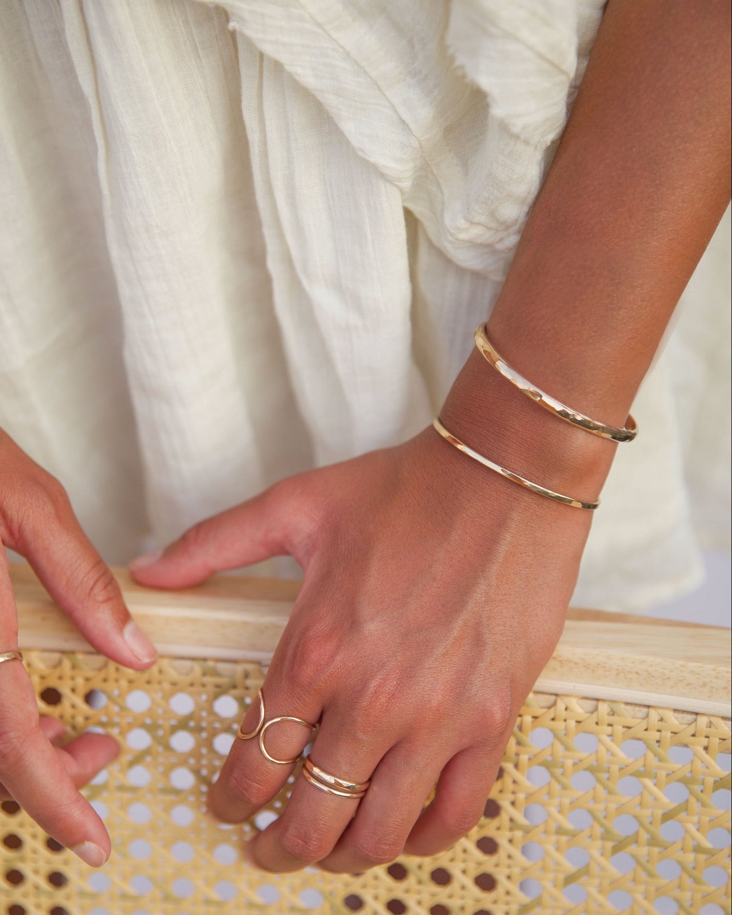 Stacked gold fill cuff bracelets worn on the wrist, styled with delicate gold rings against a light cream dress and rattan chair back.