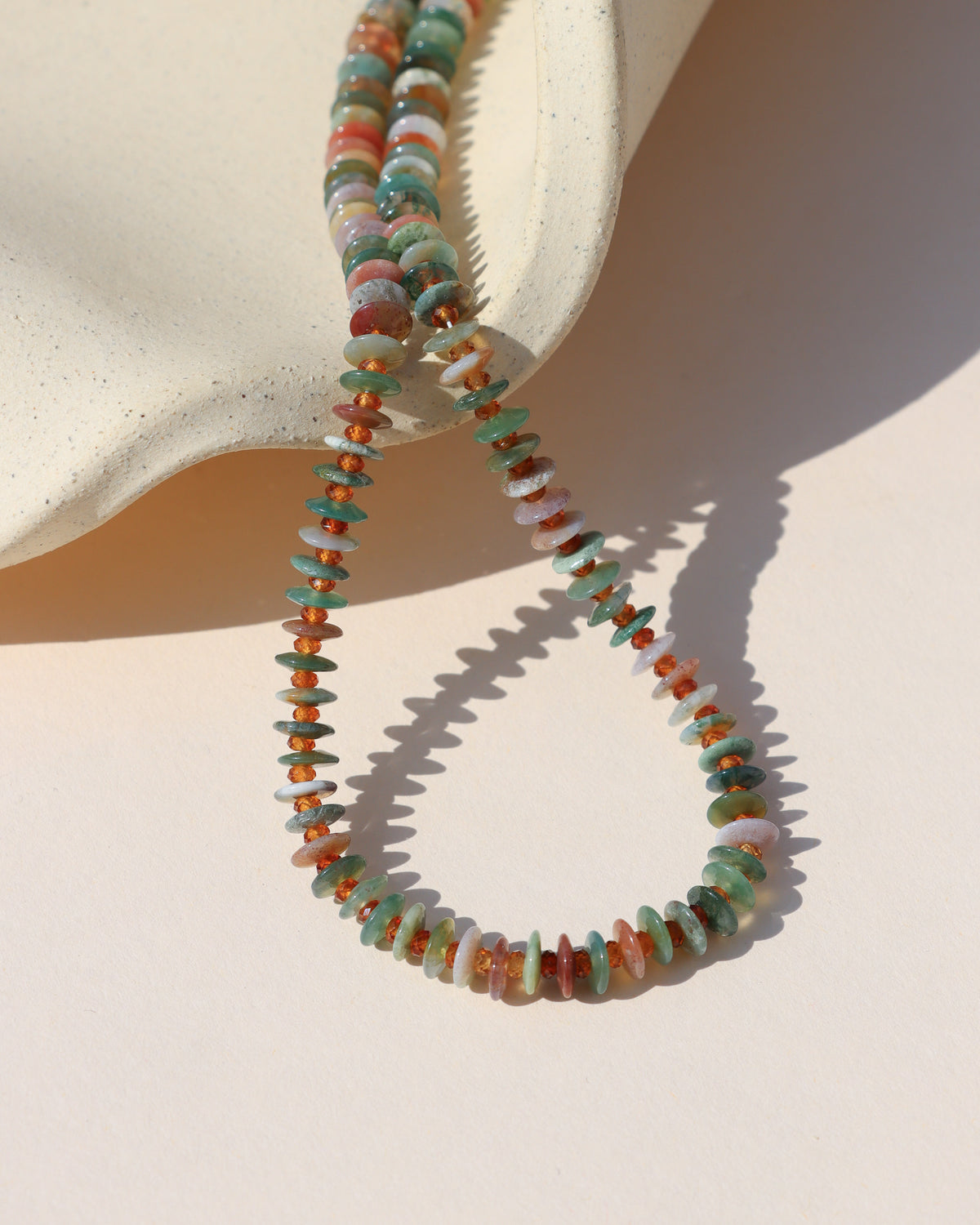 Multicolored gemstone disc bead necklace with green, turquoise, pink, cream, and amber stones, styled on a sculptural cream ceramic surface.