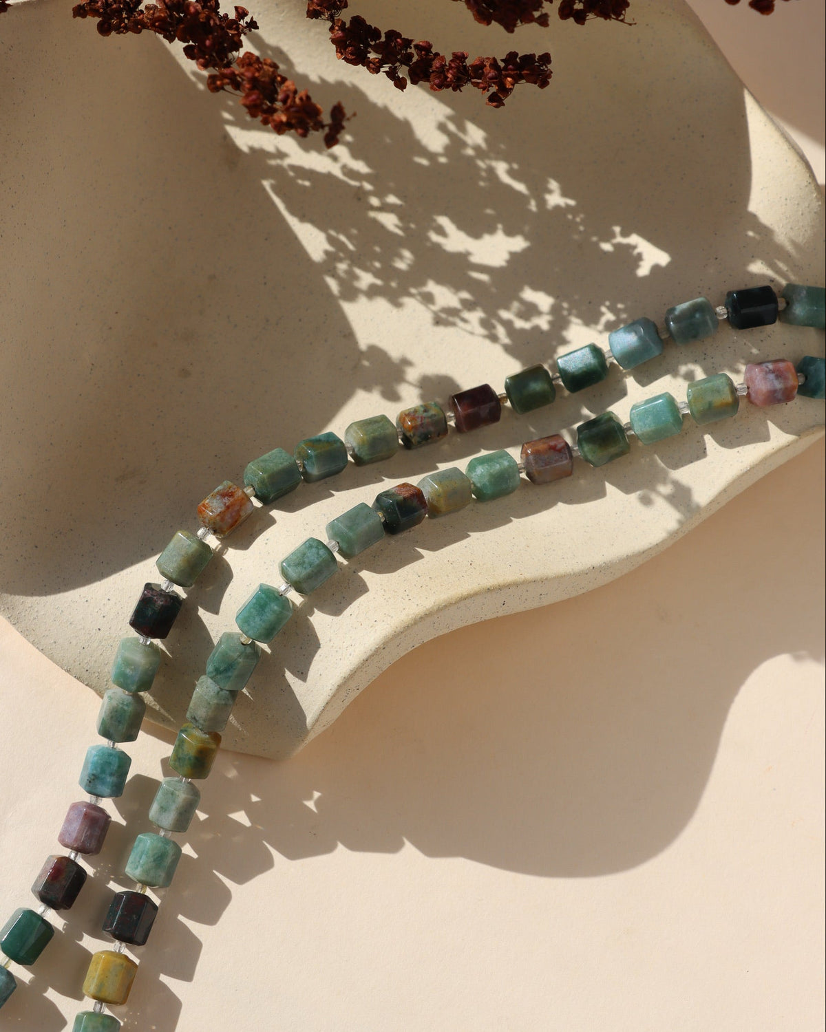 Two strands of the Fieldstone Necklace featuring multicolored Indian Agate gemstone beads in mossy green, teal, earthy brown, and warm mineral tones, draped across a textured cream ceramic dish in natural sunlight.