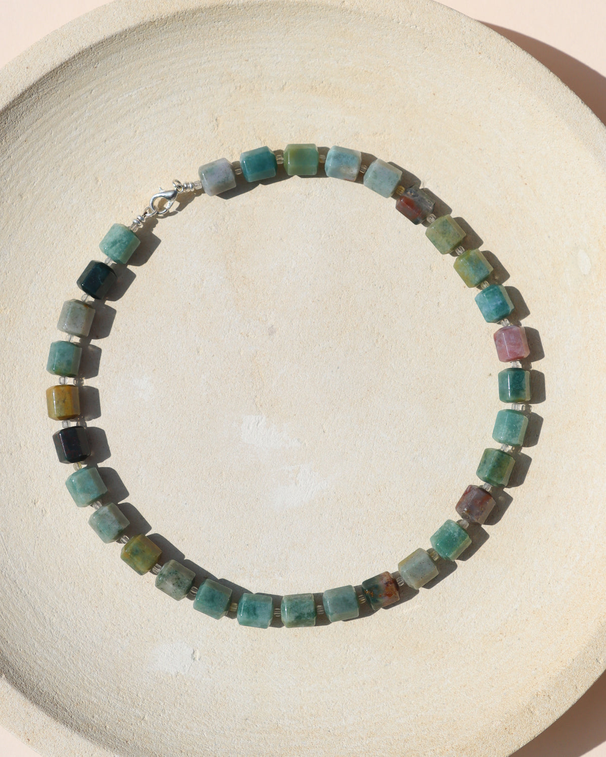 Chunky multicolored stone bead necklace with square beads in turquoise, green, blue, yellow, and earthy tones, arranged on a textured cream ceramic surface.