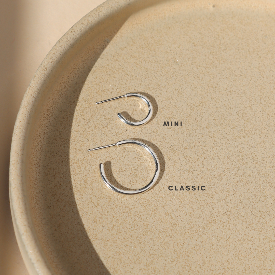 Two polished silver open hoop earrings in mini and classic sizes, displayed on a textured cream ceramic tray in warm sunlight to show the difference in size.