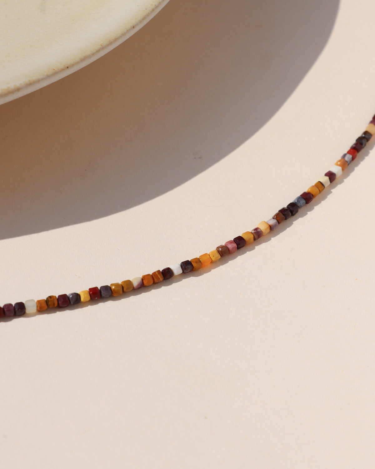 Close-up of a multicolored gemstone necklace featuring small faceted square beads in warm burgundy, orange, cream, brown, and golden tones.