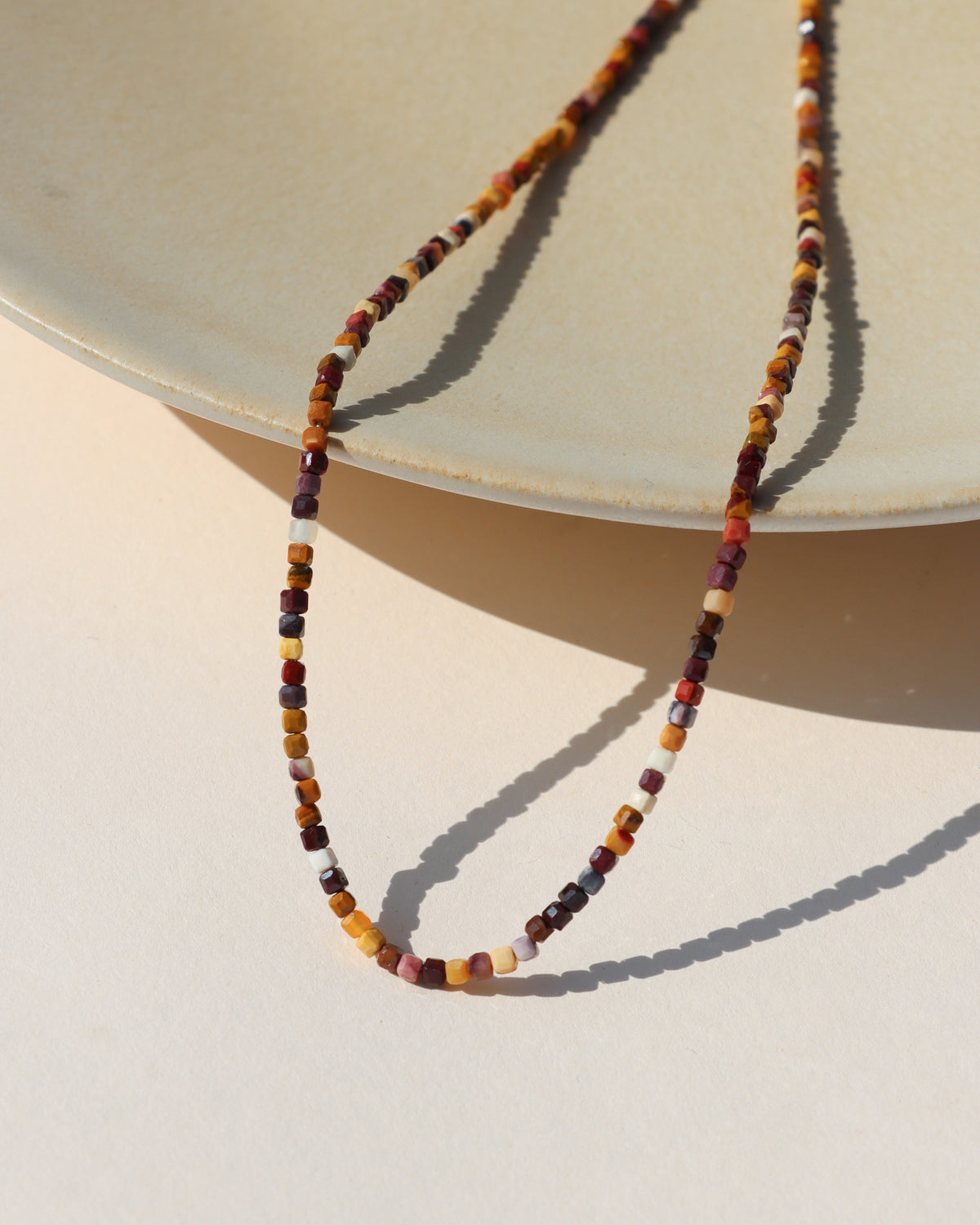 Multicolored gemstone beaded necklace with tiny faceted square beads in burgundy, rust, mustard, cream, and earthy tones, styled on a cream display in sunlight.