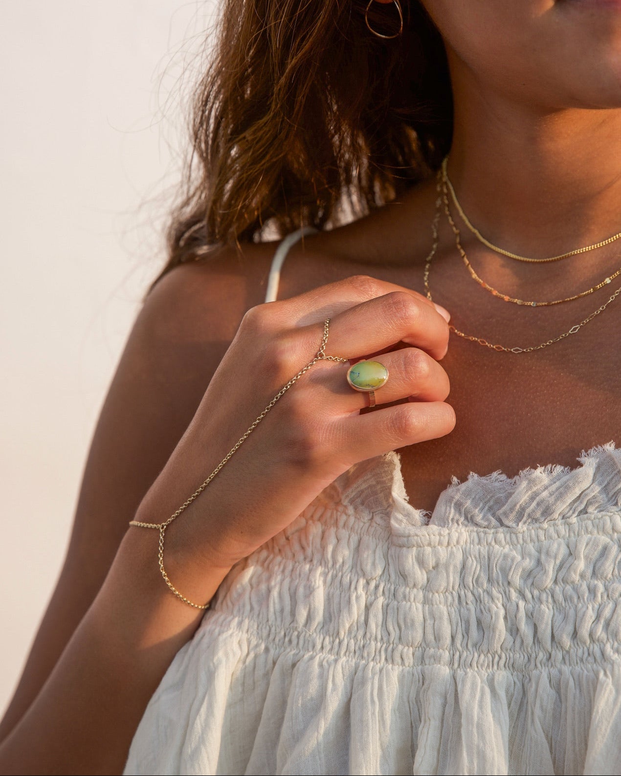 Portrait of a smiling woman in a white smocked top bathed in golden hour light, styling delicate gold jewelry: three stacked fine chain necklaces, open teardrop hoop earrings, a fine gold hand chain ring-bracelet, and an oval green turquoise gemstone ring on her middle finger.