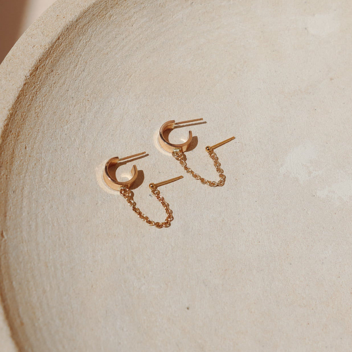 A 14k gold fill hoop is connected to a gold  classic stud earring by a rolo gold chain on a beige background in the sunlight
