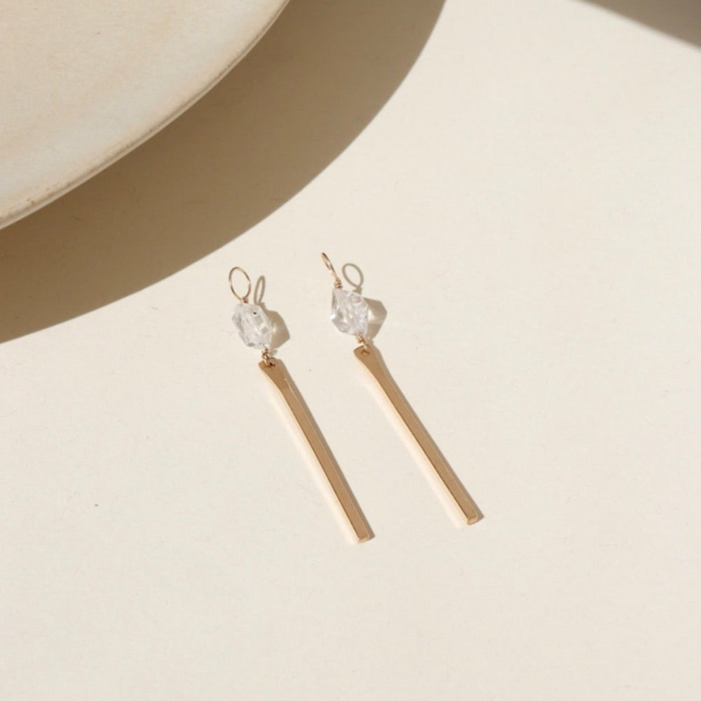 14k gold fill drop hoop charms with a herkimer gemstone wire wrapped to a gold drop post. The earrings are displayed on a cream backdrop in the sunlight 