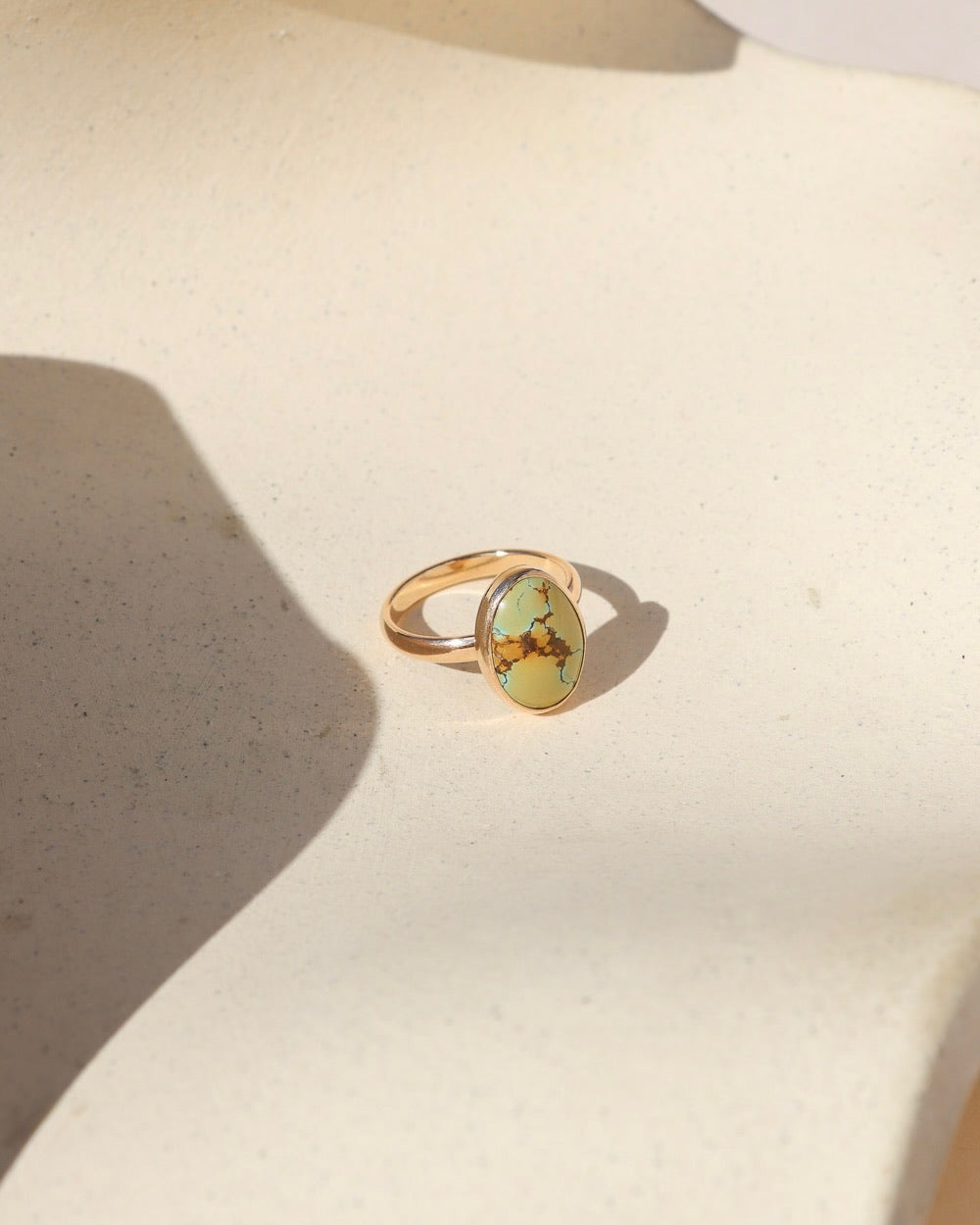Oval turquoise gemstone ring with brown veining and a gold band, displayed in warm sunlight on a neutral surface.