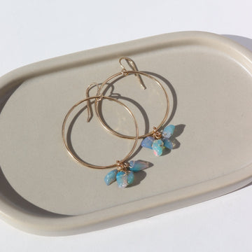 14k gold fill Rainbow Hoops laid on a gray plate in the sunlight. These earrings feature a hammered hoop with a hook earring along with a cluster of Opal pebbles.