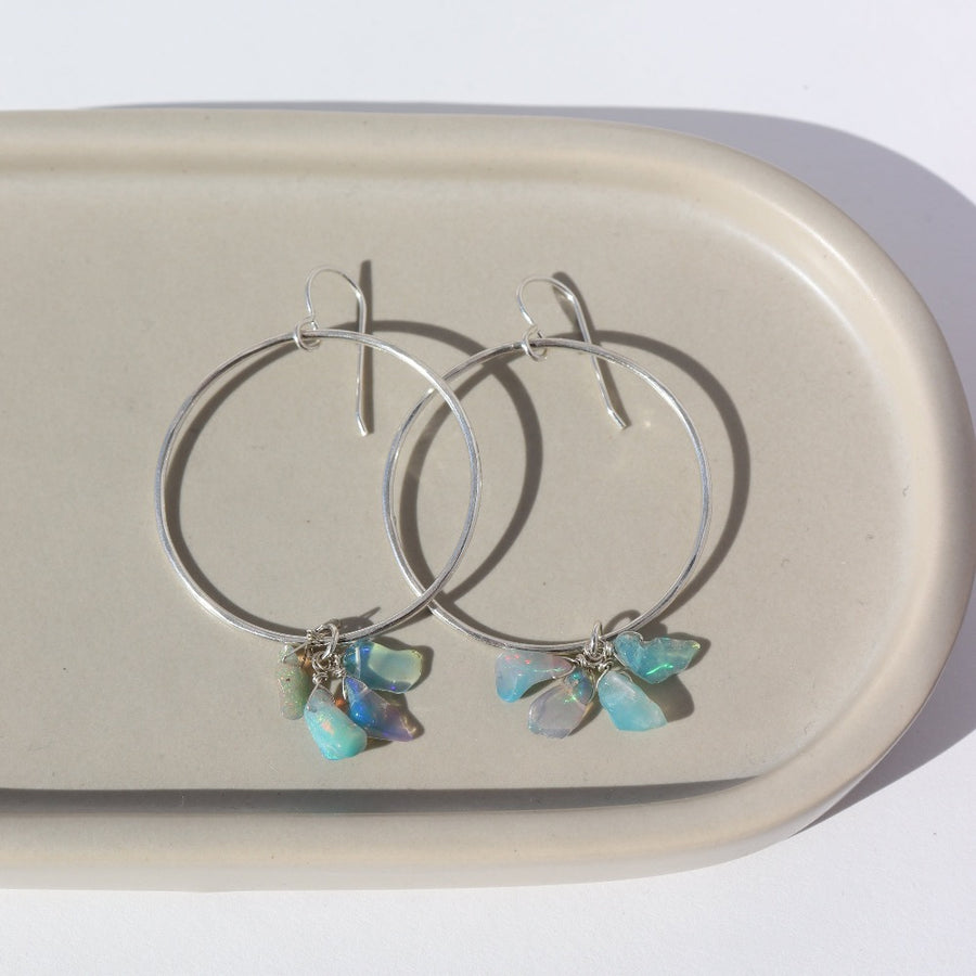 925 Sterling silver Rainbow hoops laid on a gray. These earrings feature a hammered hoop with a hook earring along with a cluster of Opal pebbles.