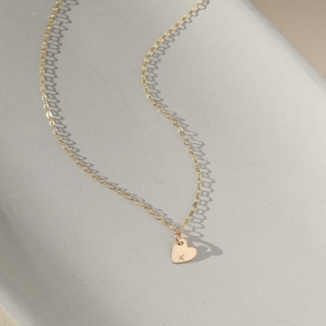 a dainty heart charm with an initial stamped on them, on a necklace chain, photographed on a ceramic dish