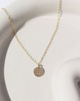 14k gold fill Tiny Monogram Disc Necklace laid on a white plate in the sunlight. This necklace features our simple chain with a disc pendent with a letter stamped on it.