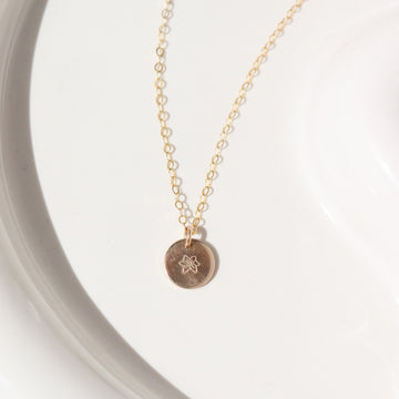 14k gold fill Tiny Birth Flower Disc Necklace laid on a white plate in the sunlight. This necklace features our simple chain with a birth flower pendent of your choice.