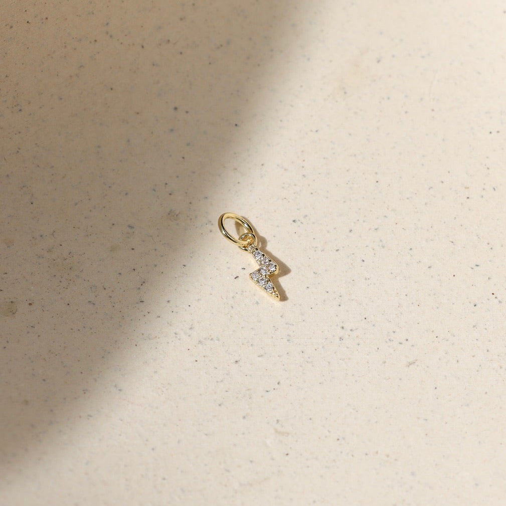 Gold lightning bolt charm with pavé crystal details, photographed on a warm neutral surface with soft natural light and shadows.