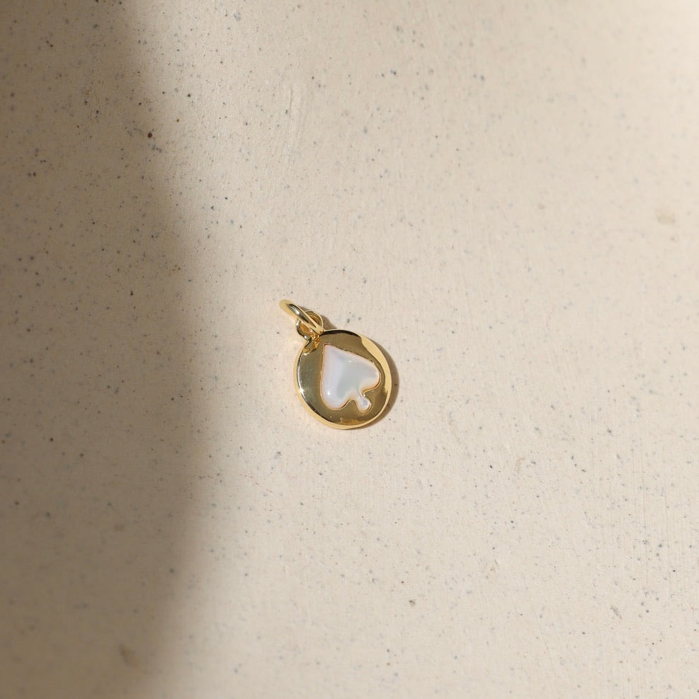 Gold coin charm featuring a mother of pearl spade inlay, photographed on a warm neutral surface with soft natural light and shadows.