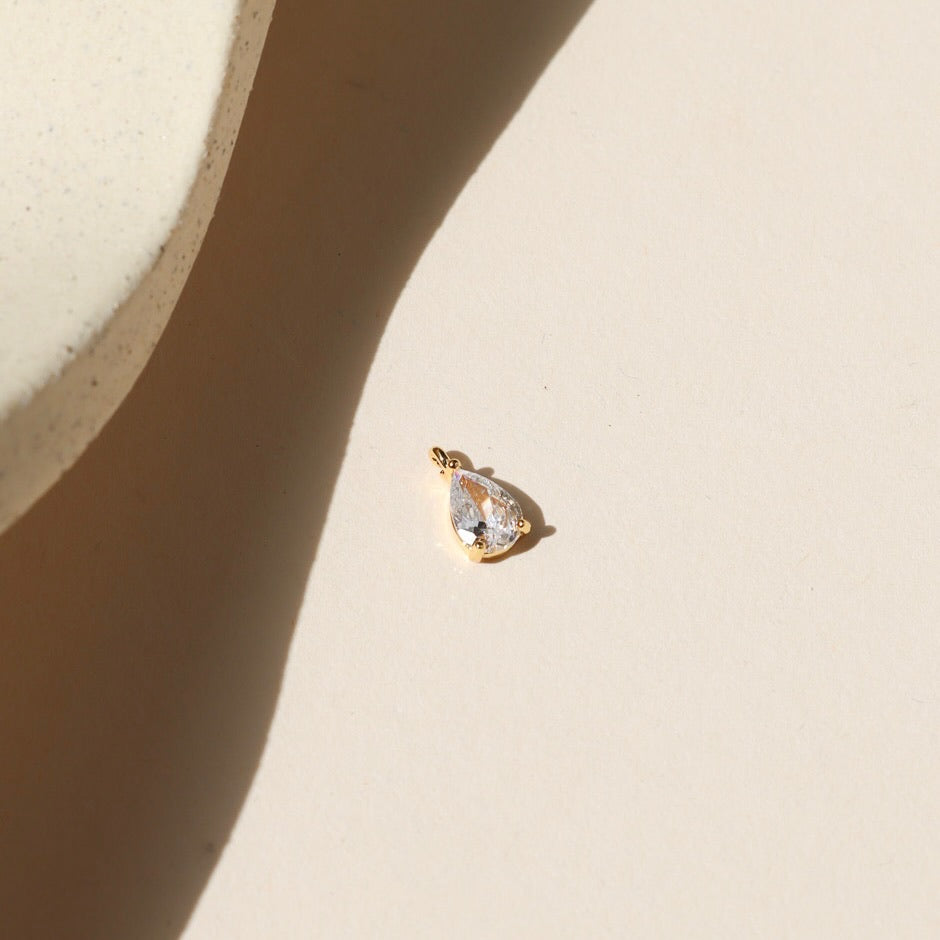 Gold charm featuring a clear teardrop-cut crystal, photographed on a warm neutral surface with soft natural light and shadows.