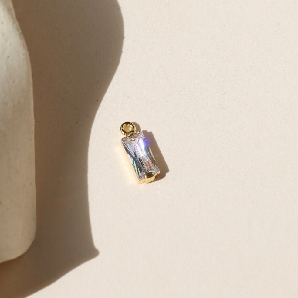 Gold charm featuring a clear baguette-cut crystal, photographed on a warm neutral surface with soft natural light and shadows.