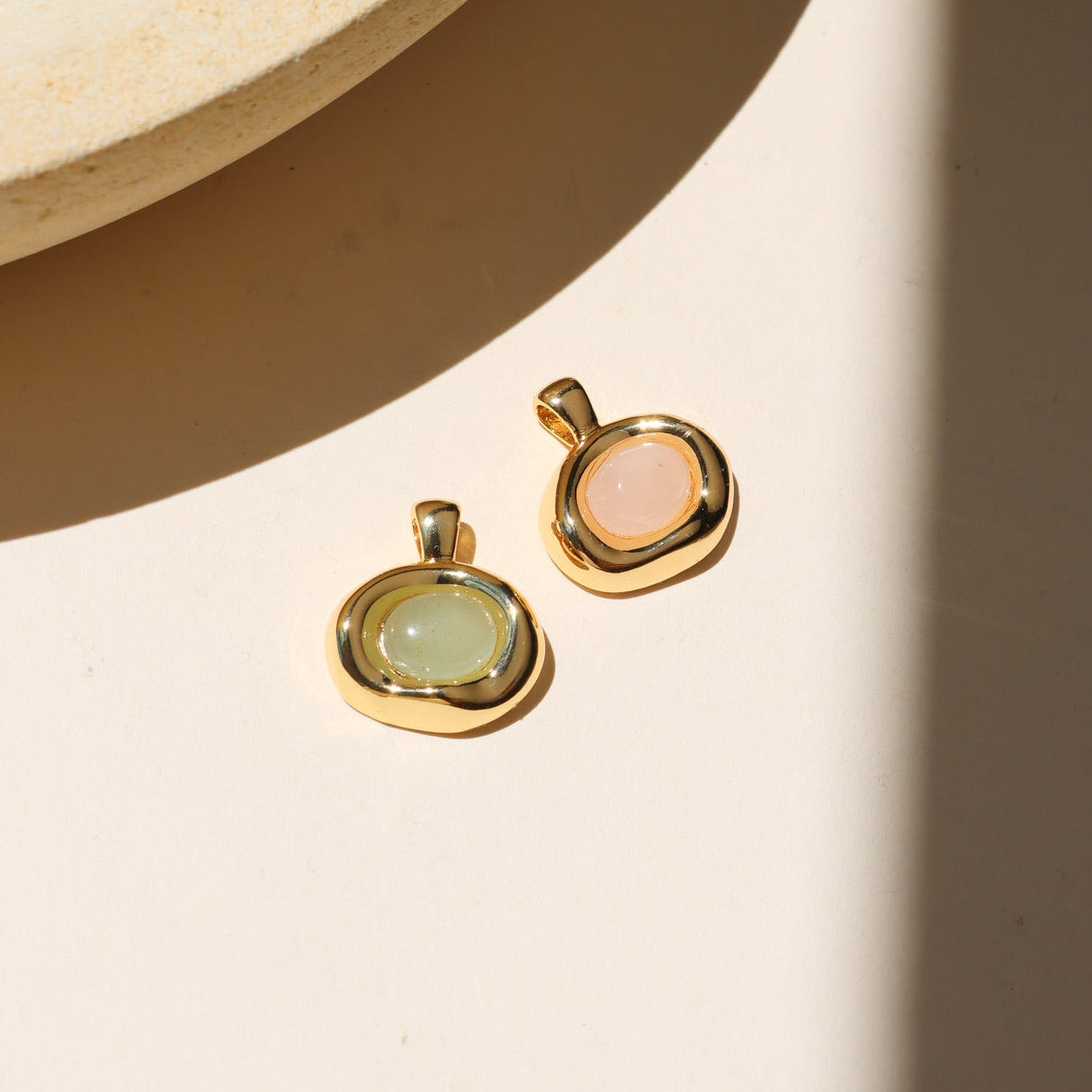 Gold pendant charms featuring aventurine and rose quartz cabochons in organic gold settings, displayed on a neutral surface in warm natural light.