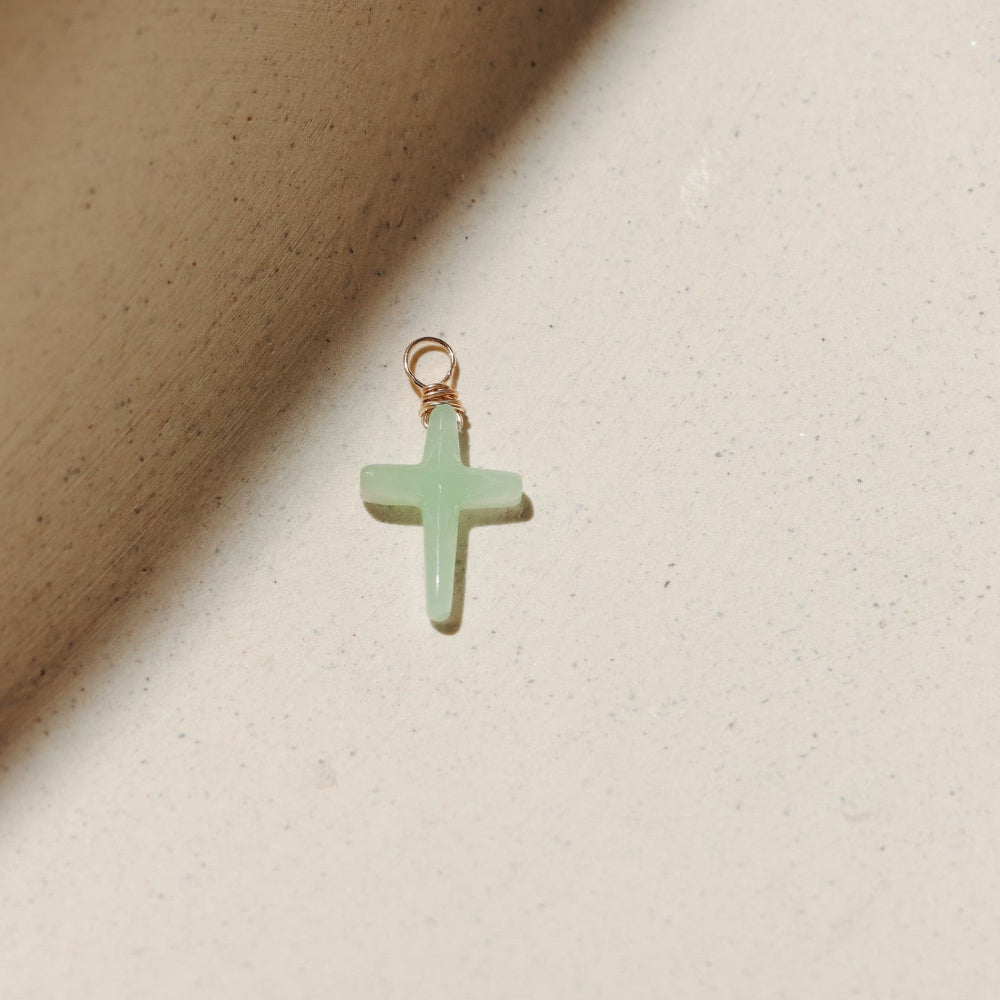 Chrysoprase cross charm featuring a soft green gemstone cross wrapped with gold wire, photographed on a warm neutral surface with soft natural light and shadows.