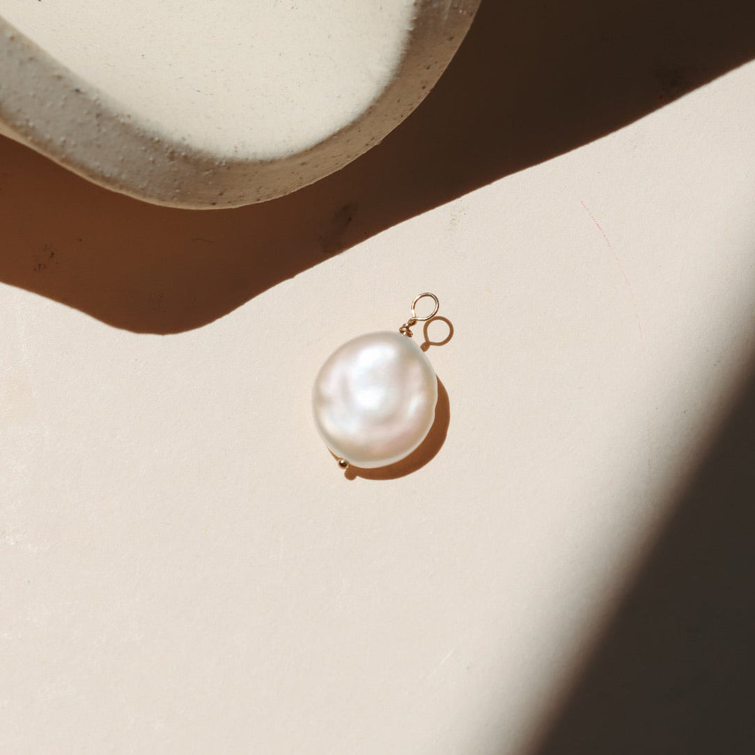 Freshwater pearl charm with a naturally irregular pearl suspended from a gold loop, photographed on a warm neutral surface with soft natural light and shadows.