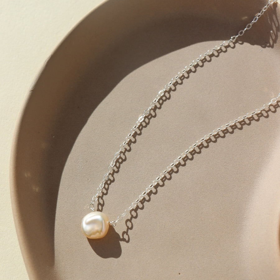 925 sterling silver freshwater pearl necklace laid on a peach colored plate. This necklace features the simple chain with the freshwater pearl wired in the middle.